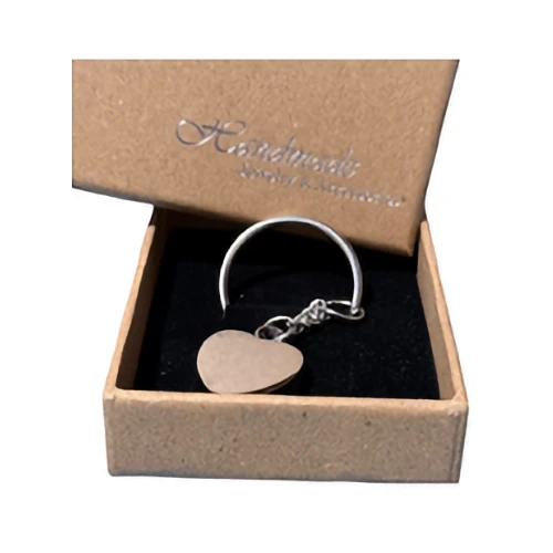 A heart-shaped silver charm, suitable for a keepsake or memorial, is presented inside an open, kraft paper gift box with a black interior.